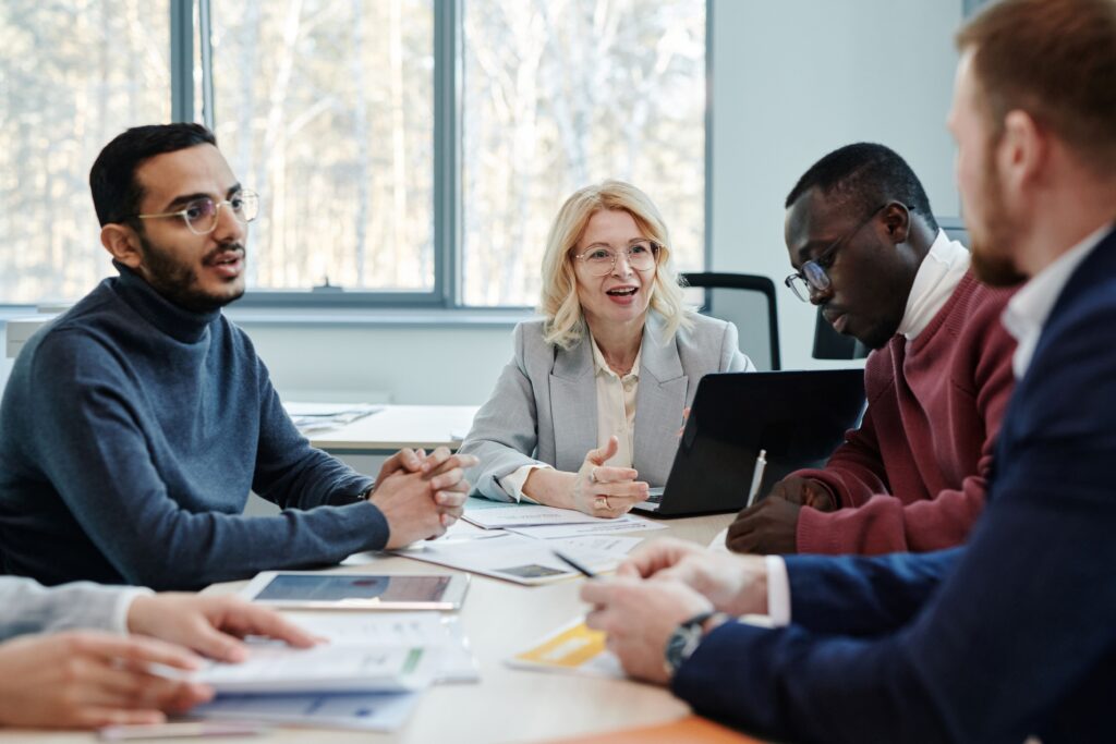 Five people around a meeting table in conversation, with printed charts and a laptop.