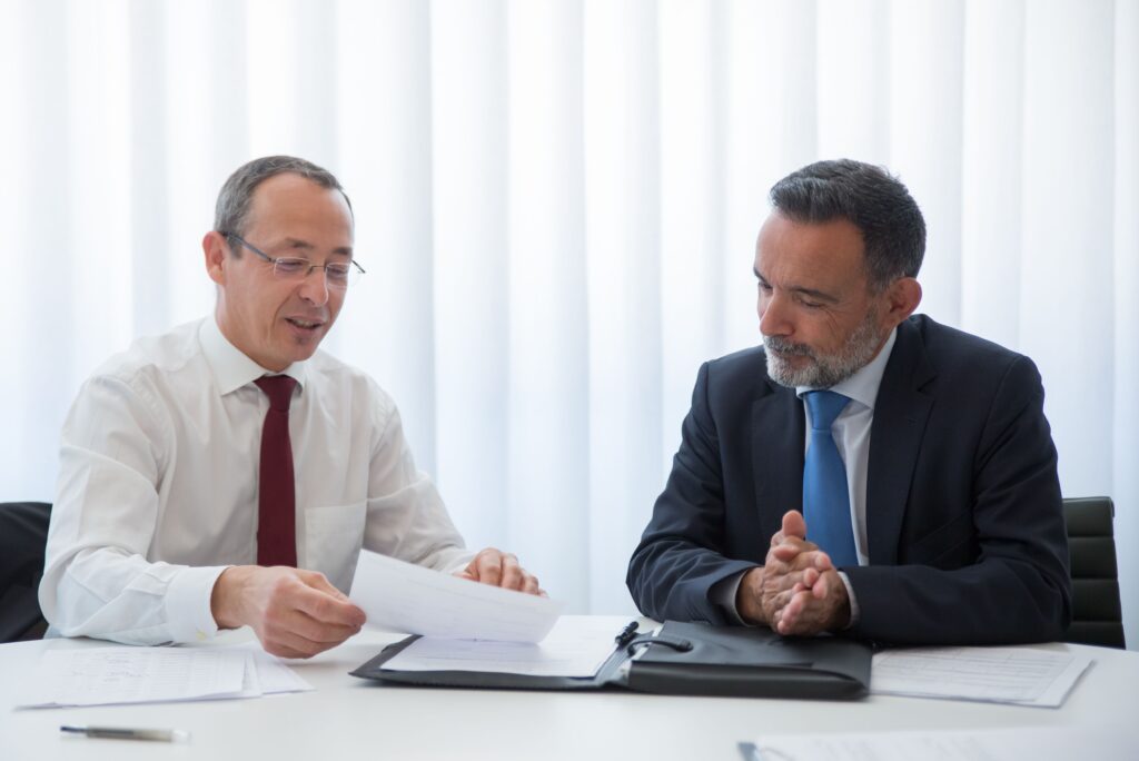 Two people side by side at a desk, one reading from a stapled document in a folder.
